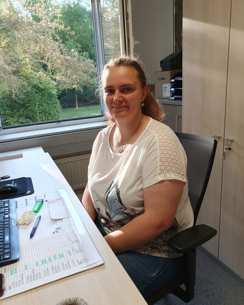 Eine Mitarbeiterin sitzt an ihrem Arbeitsplatz in einem hellen Büro. Auf dem Schreibtisch liegen Tastatur, Unterlagen und Arbeitsmaterialien, im Hintergrund ist eine grüne Außenanlage zu sehen.
